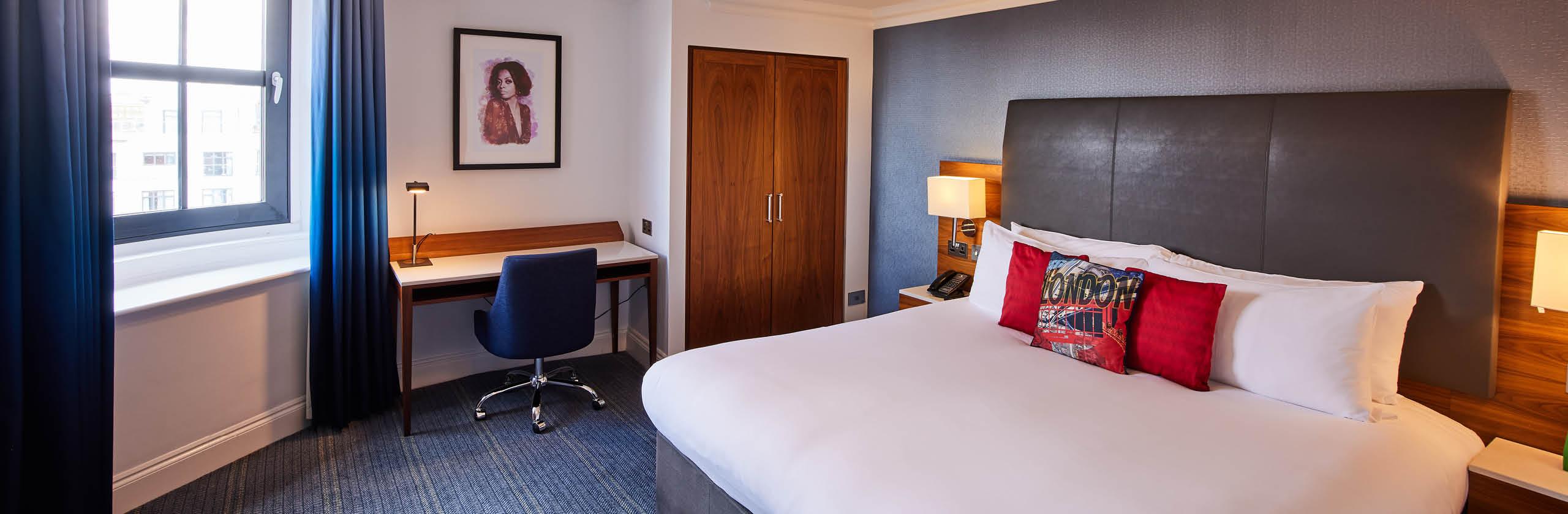 Modern hotel bedroom with a large double bed, grey upholstered headboard, red accent cushions, a work desk with chair by the window, wooden wardrobe, and soft lighting
