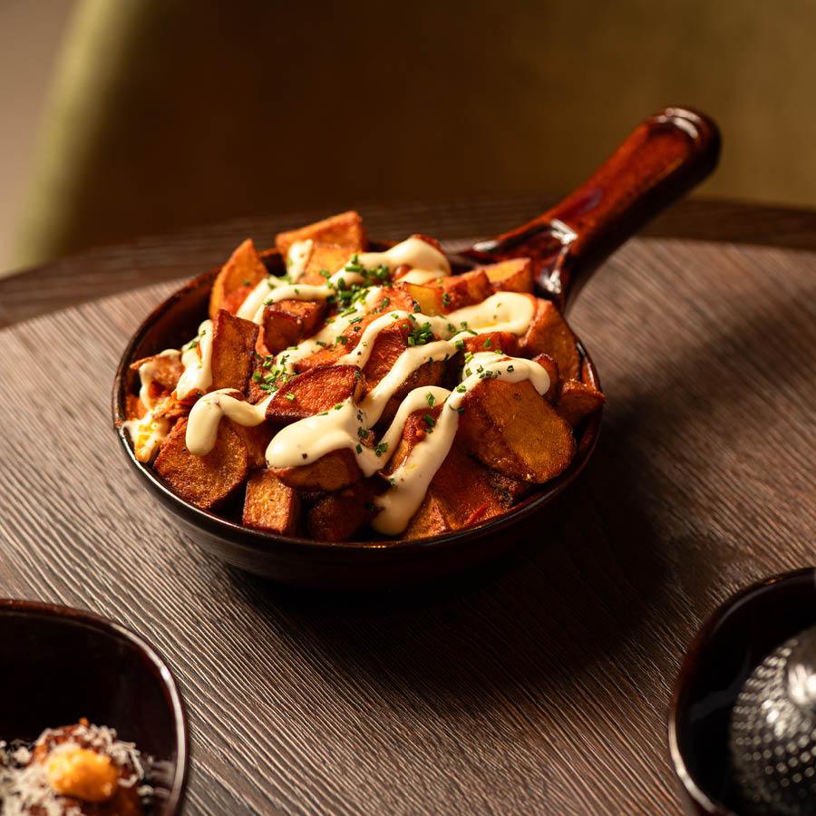 Crispy roasted potatoes served in a small pan, topped with creamy sauce and garnished with chopped herbs on a wooden table.
