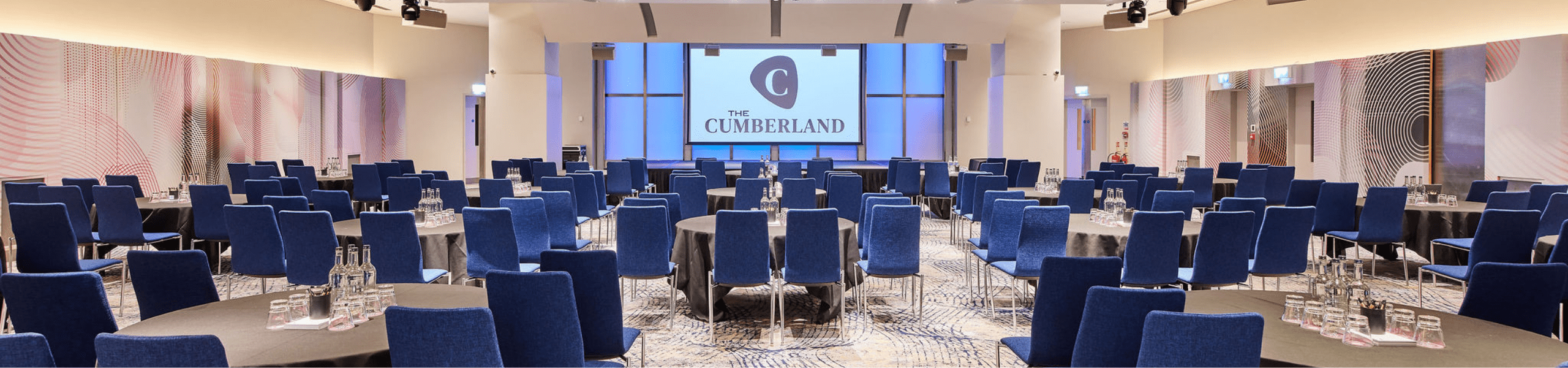 Conference and events space at The Cumberland hotel, arranged in cabaret style with round tables, blue chairs, bottled water, and a large presentation screen displaying The Cumberland logo.