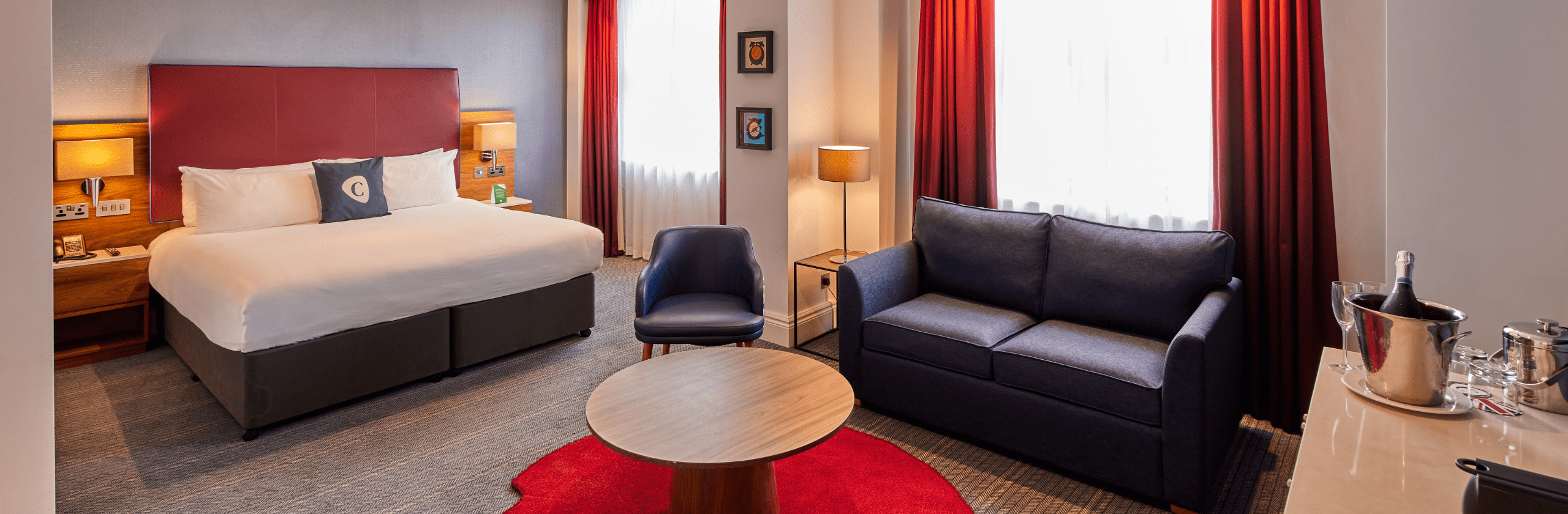 Spacious hotel room featuring a king-size bed with a red upholstered headboard, a blue sofa and armchair, round coffee table, floor lamp, large windows with red curtains, and a drinks station with a bottle of sparkling wine.
