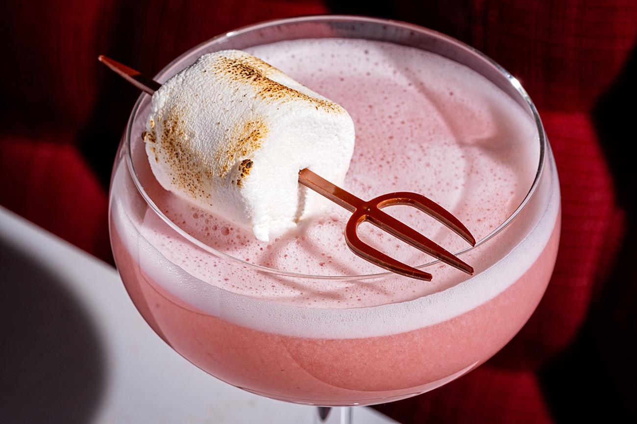 A pink cocktail with Hell's kitchen shaped cocktail stick and a fluffy marshmallow