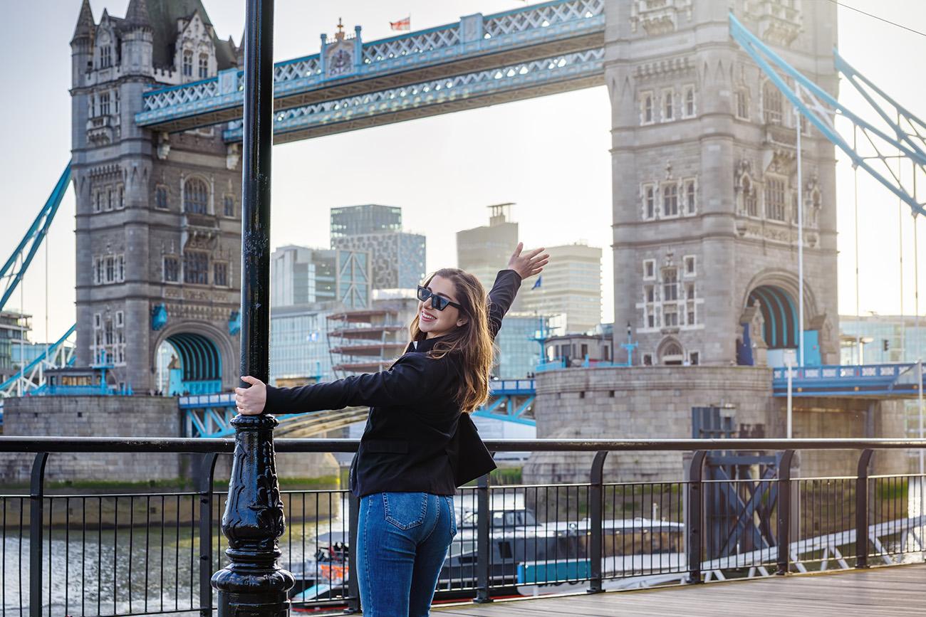 An excited, young woman stands in front of the Tower Bridge in London