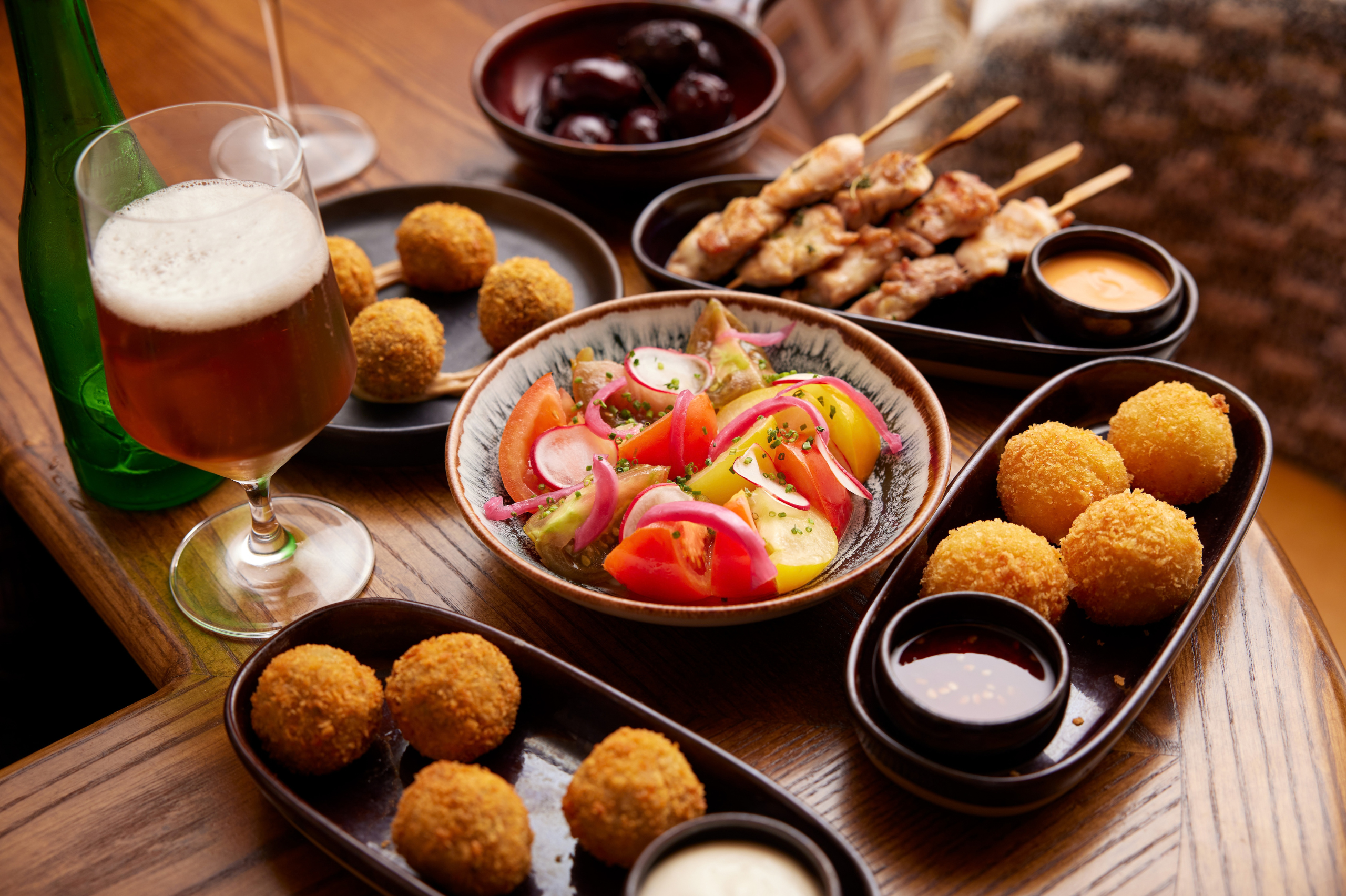 Selection of sharing plates and drinks on a wooden table, featuring a fresh tomato salad, crispy croquettes, grilled skewers, dipping sauces and a glass of beer.