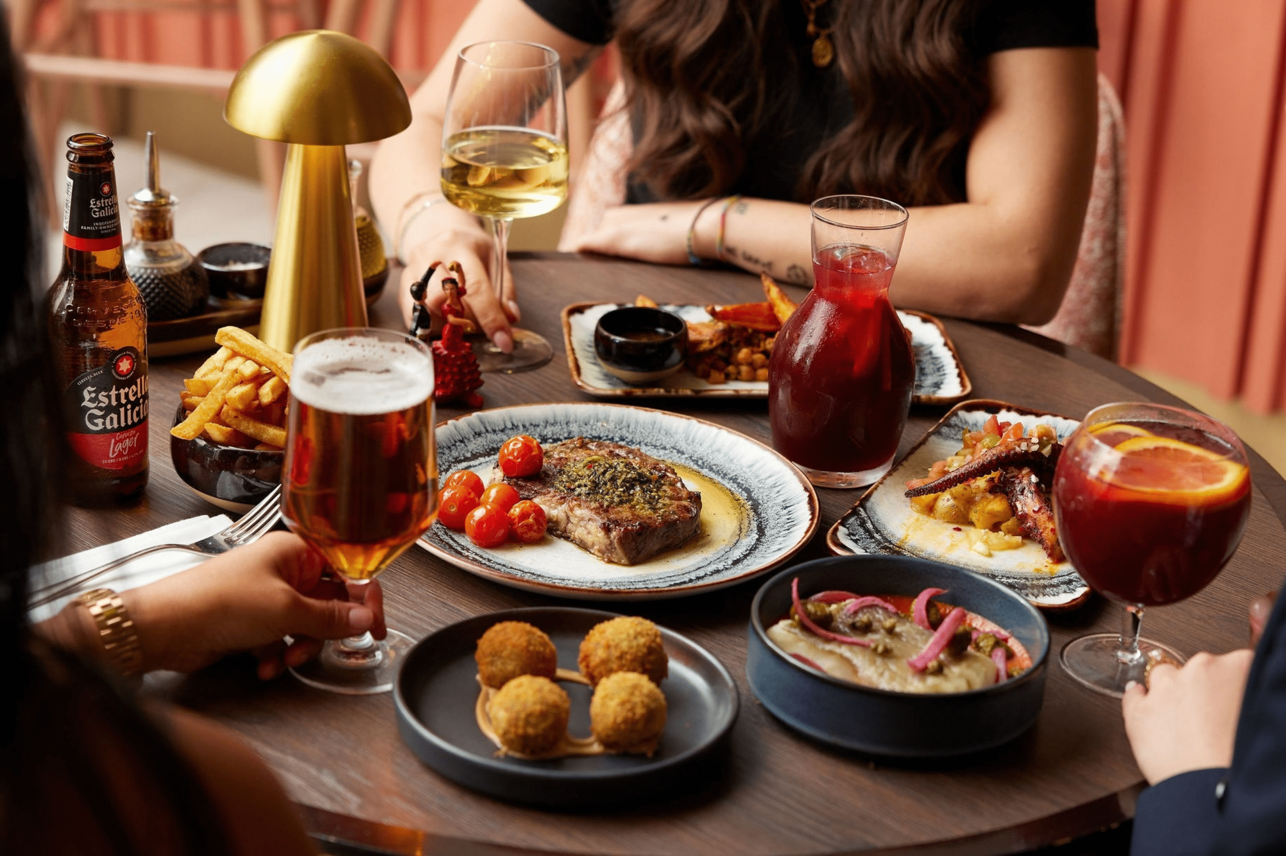 Selection of Spanish-inspired dishes and drinks shared around a restaurant table, featuring steak, croquettes, tapas-style plates, sangria, beer and wine in a warm dining setting.