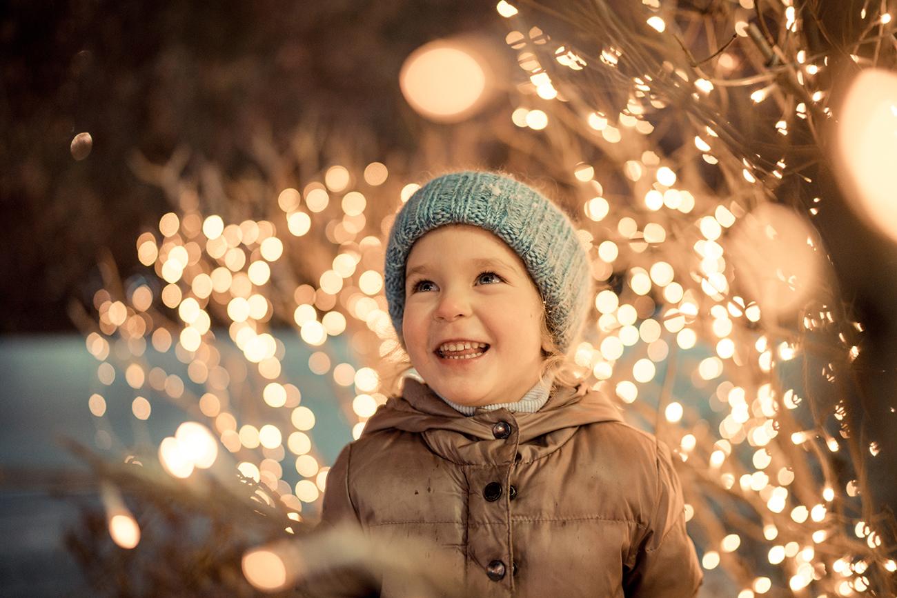 Smiling child in a blue knitted hat and winter coat admiring golden fairy lights wrapped around tree branches.