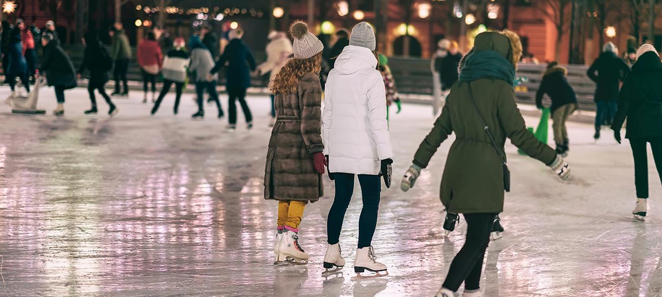 People in winter coats and knitted hats skating on an outdoor ice rink beneath festive lights at night