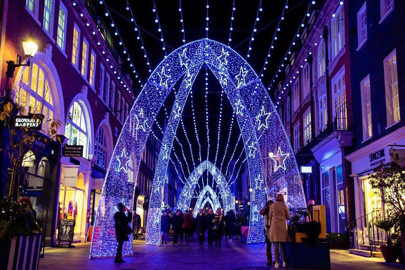 Shoppers walking beneath arches of blue Christmas lights decorated with white stars along a London shopping street at night.
