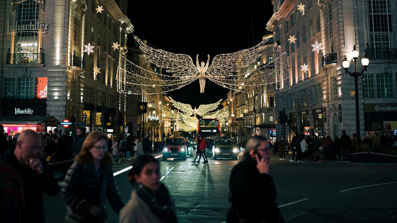 Golden angel Christmas lights suspended above Regent Street at night, with shoppers, taxis and a red double-decker bus below.